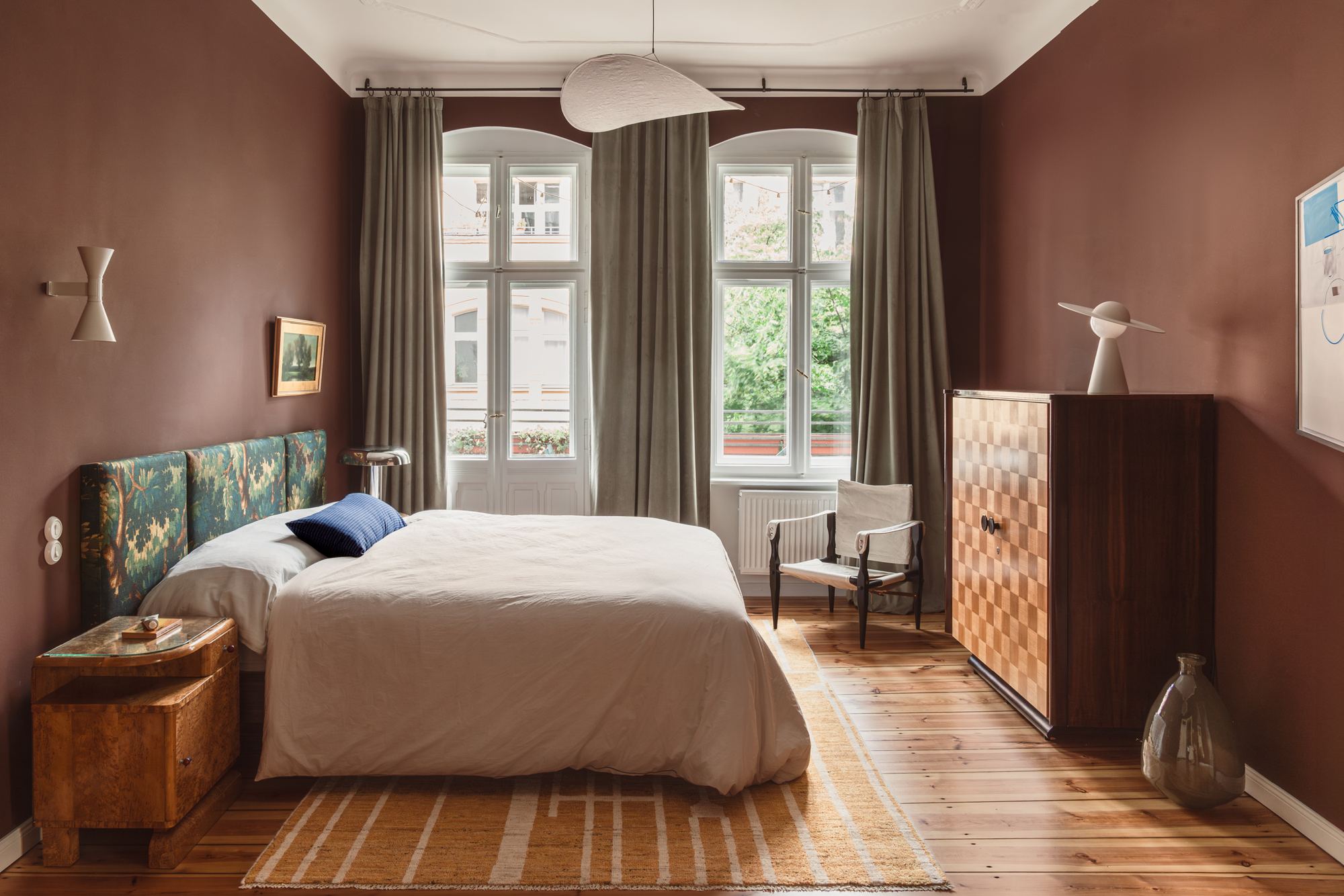 The bedroom, with deep oxblood walls, a king bed with a tapestry headboard and two tall windows behind it.