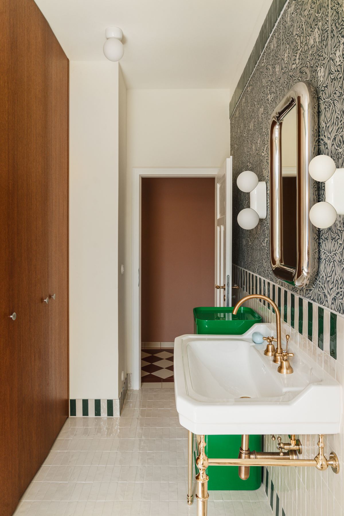 The full length of the bathroom, with a tall wooden cupboard on the left, the sink and mirror on the right, and the checkerboard hall floor visible through the doorway.