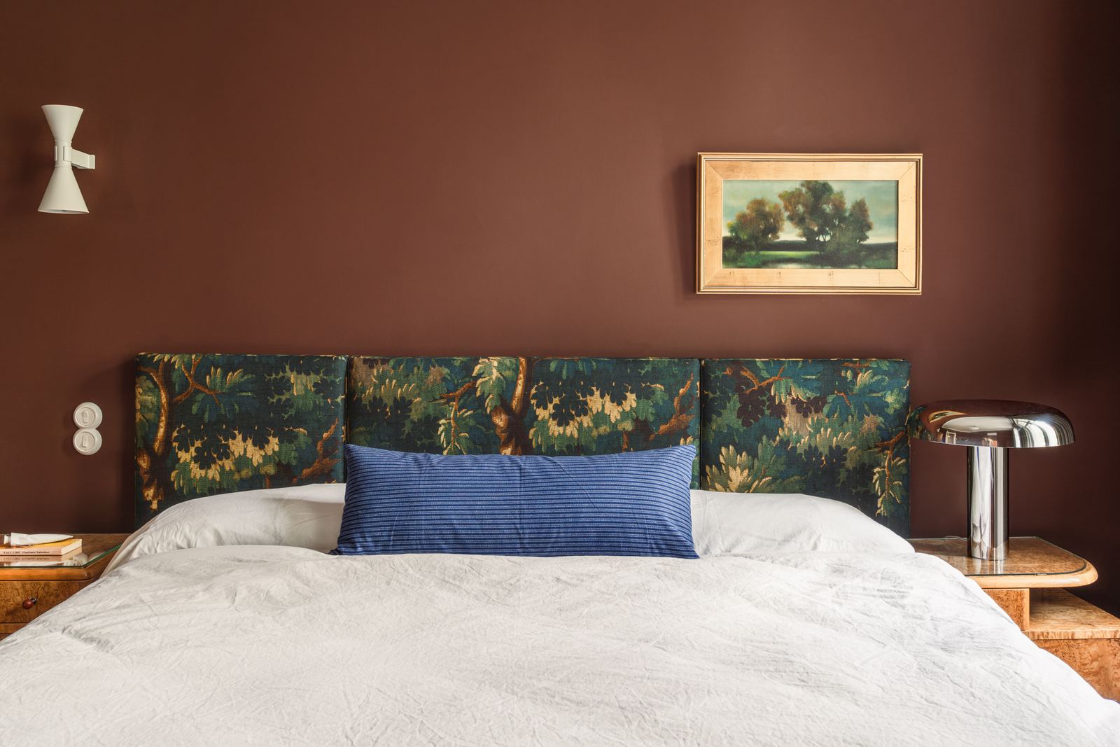 A king bed made up in white linen with a blue bolster, against a headboard upholstered in green and blue tapestry.