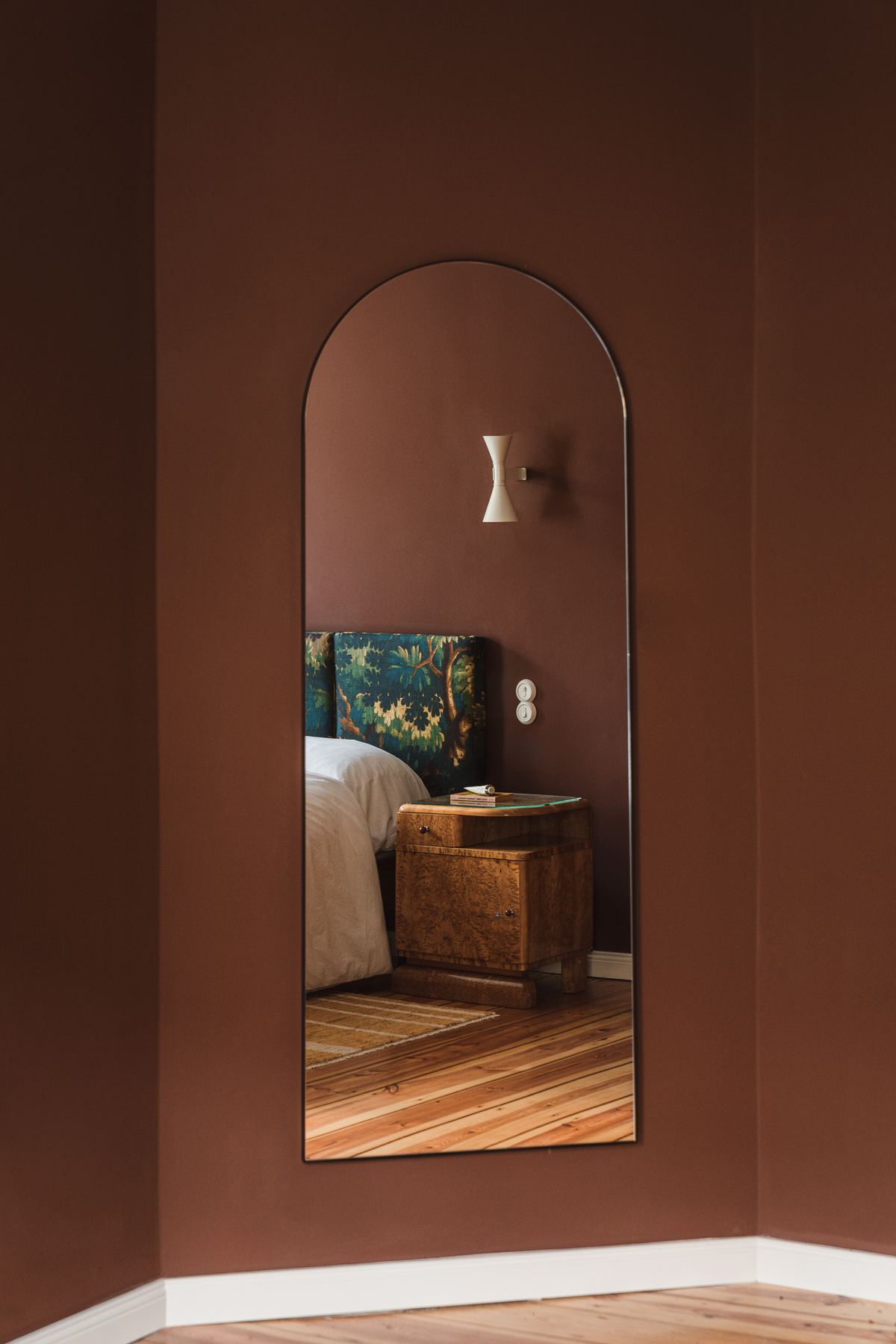 A tall arch-topped mirror on an oxblood wall, reflecting the bed and a wall light.