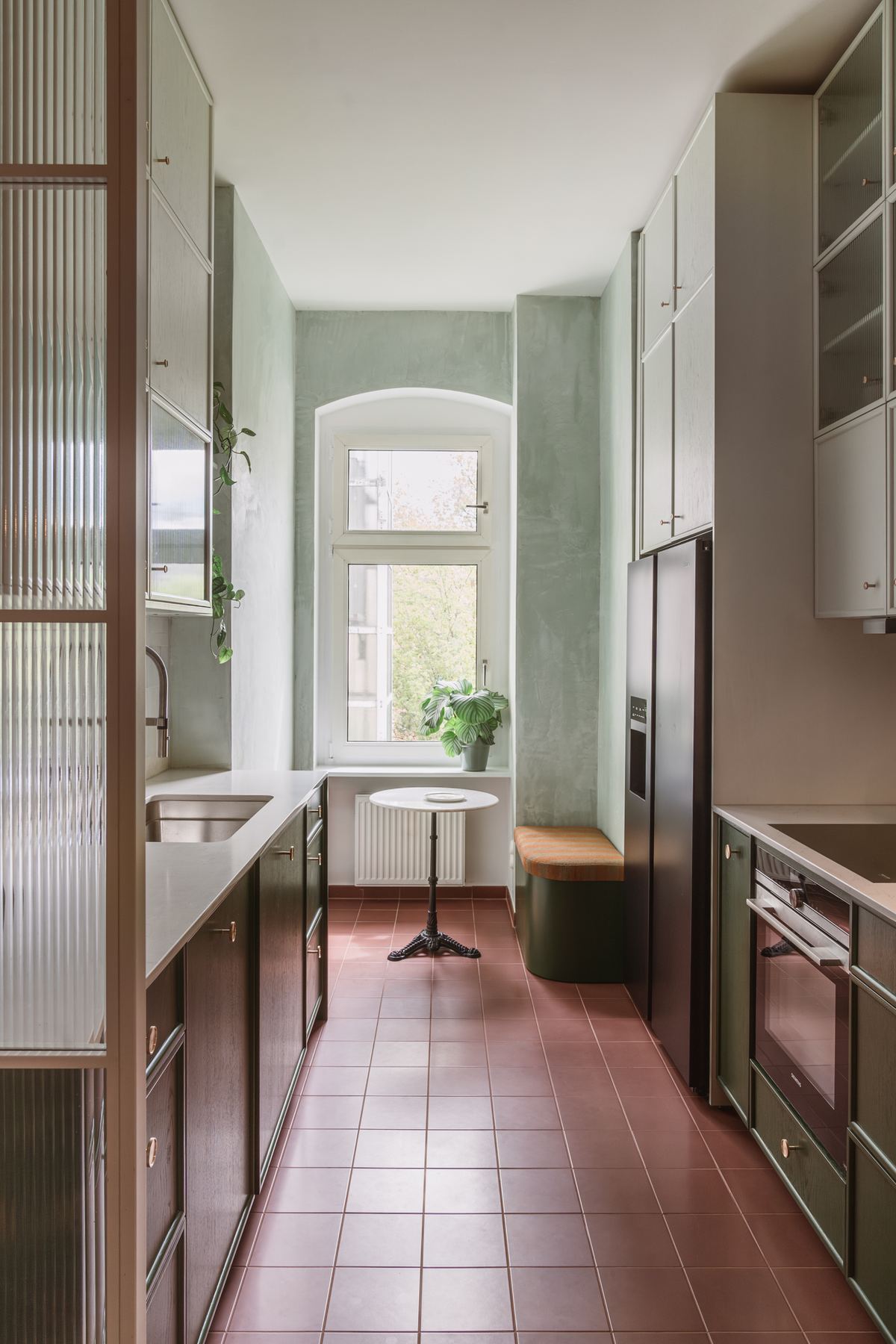 The galley kitchen with dark green cabinets, a terracotta tile floor and an arched window at the far end.