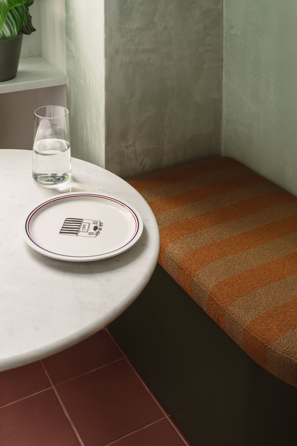 A marble-topped table beside a built-in bench upholstered in striped orange fabric, with a plate and a glass of water on the table.