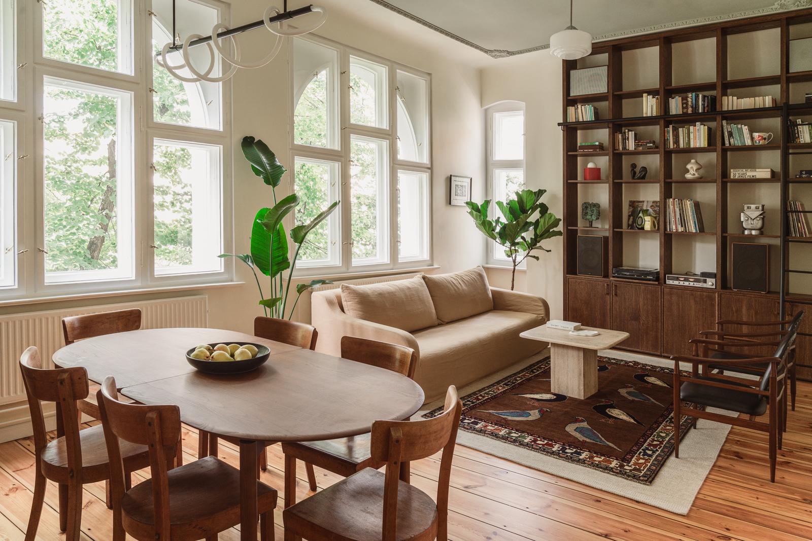 The living room with a round dining table and wooden chairs in the foreground, a sofa beyond it and tall windows along the left wall.